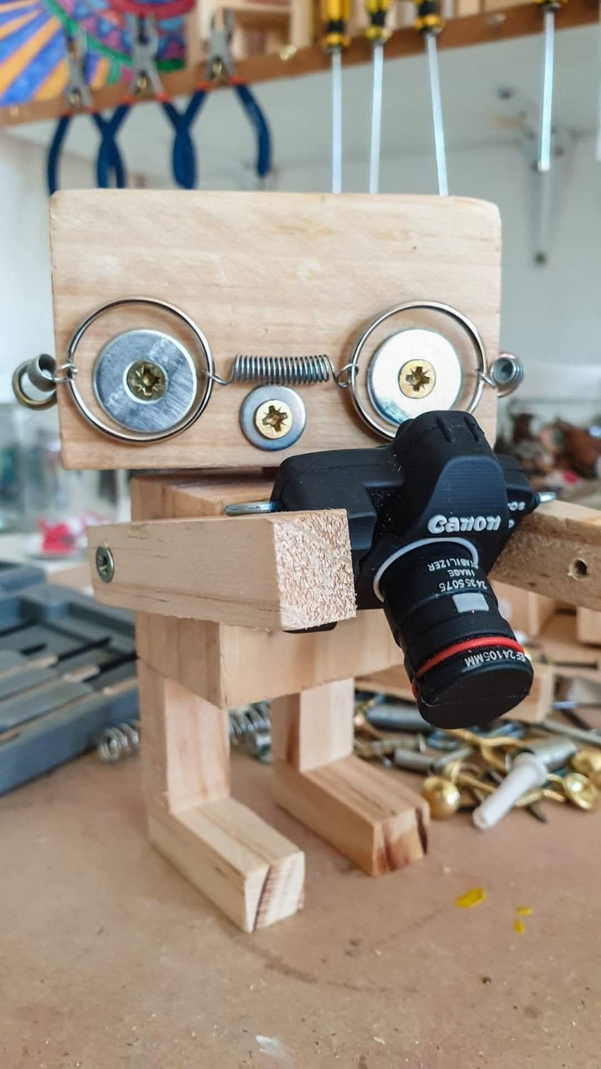 A wooden robot with washer-and-spring eyes holding a toy camera, standing on a workbench under a rack of pliers and screwdrivers