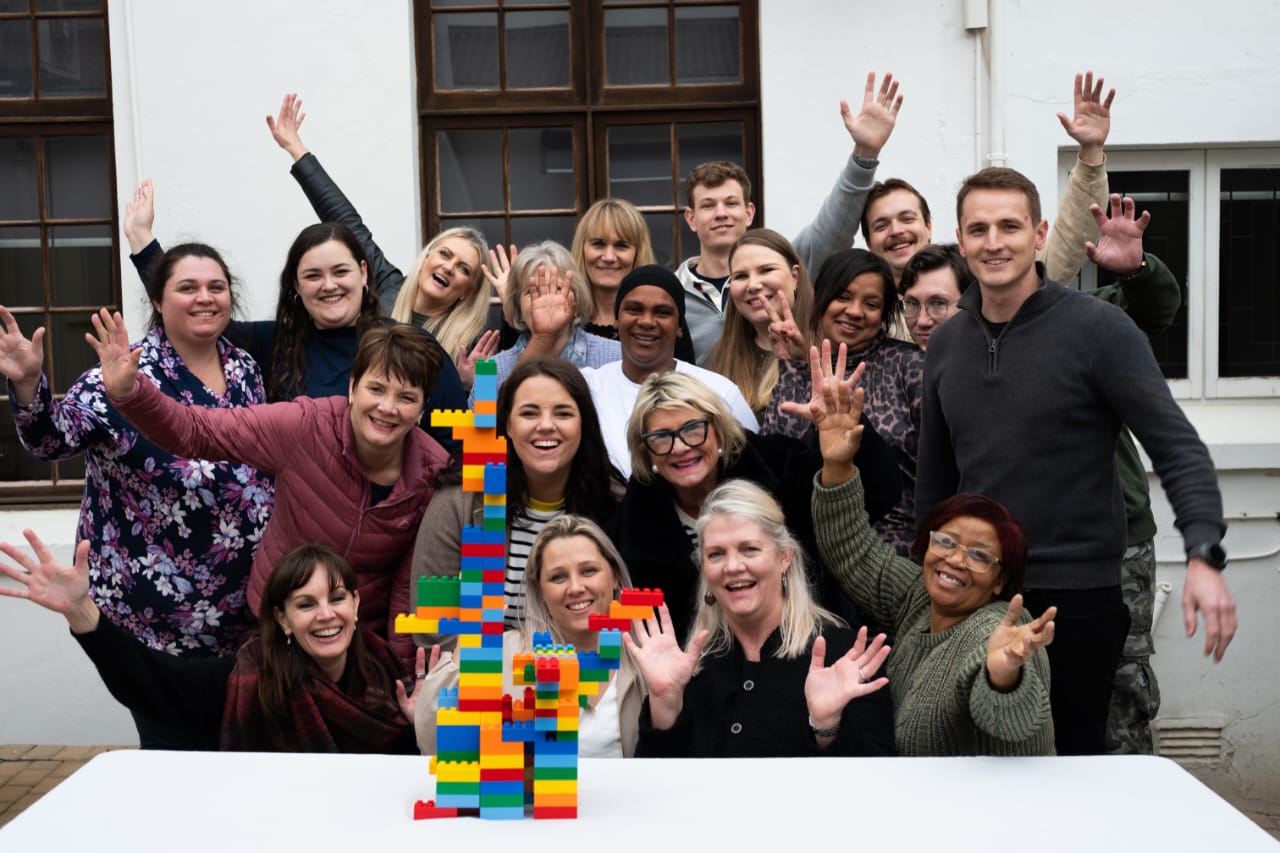 A corporate team standing together with hands raised behind a tall, colourful brick tower they built during a Brand Code Discovery day.